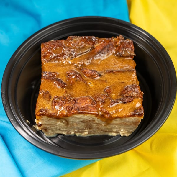 A square of bread pudding with a glossy caramel topping in a black bowl on a blue and yellow cloth.