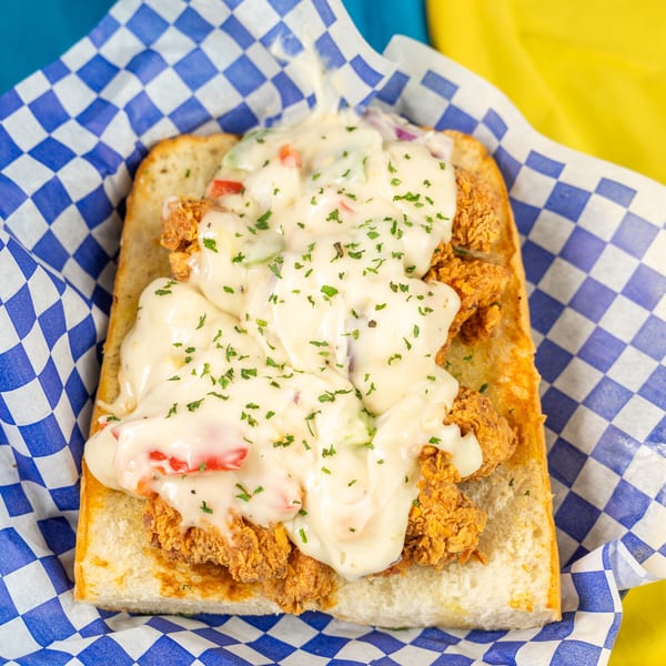 Fried chicken po'boy sandwich topped with creamy sauce, diced vegetables, and parsley in a checkered basket.