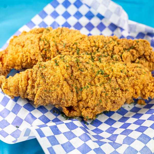 Two golden-brown fried fish fillets, sprinkled with parsley, resting in a blue and white checkered paper basket.