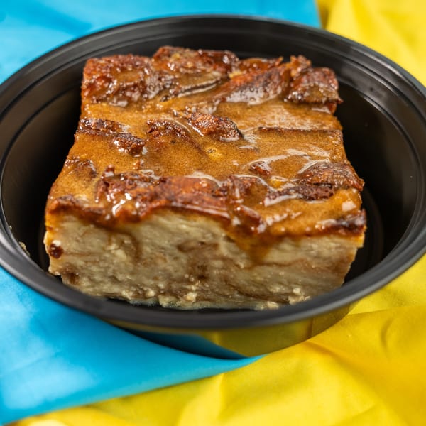 Bread pudding with caramel sauce in a black container, on a blue and yellow tablecloth.